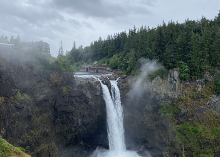 A waterfall cascades down a rocky cliff surrounded by lush green forest under a cloudy sky, creating the perfect backdrop for Seattle sightseeing. A large building is visible in the distance, offering an ideal spot for private Seattle tours.