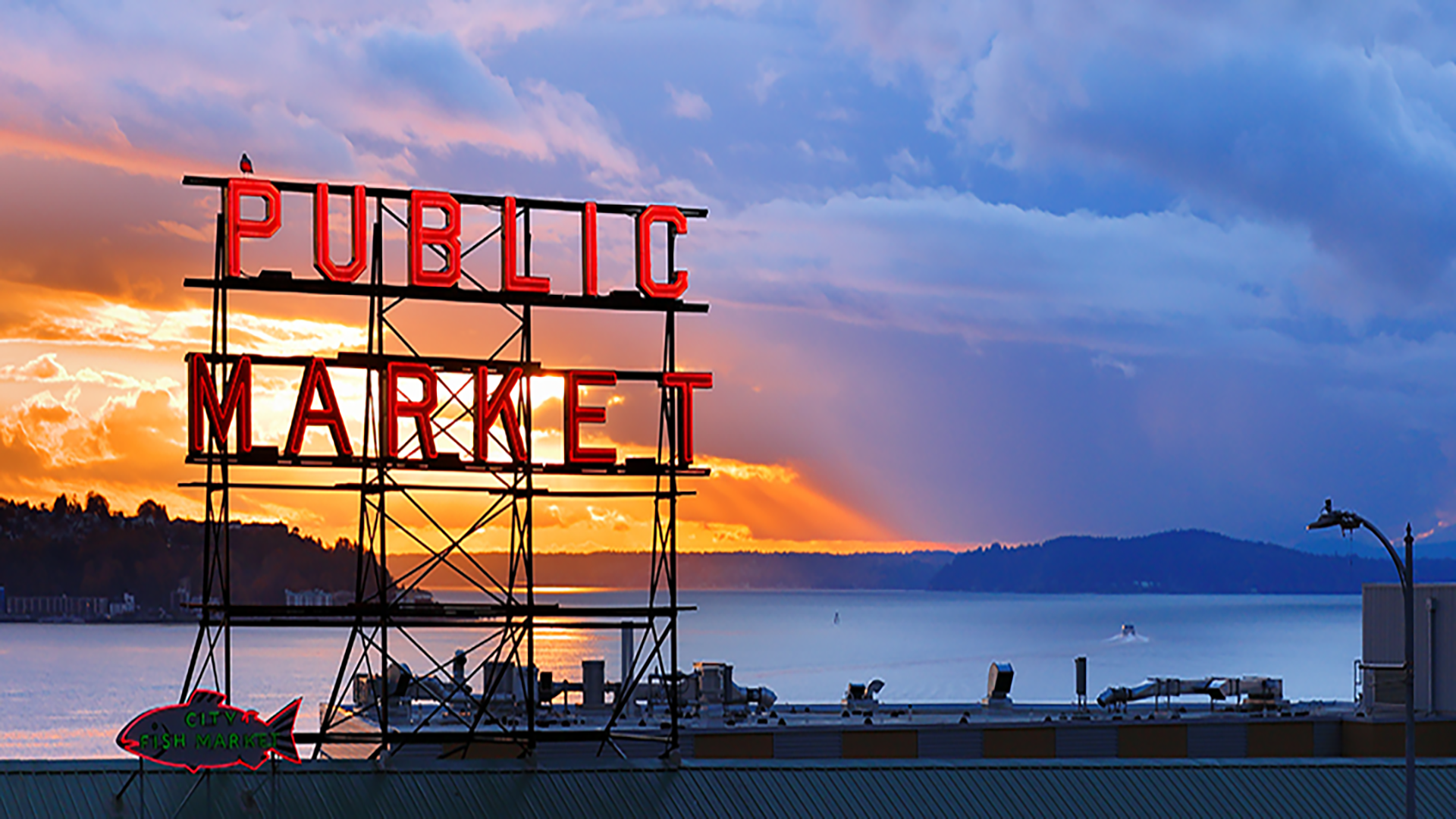 A neon "PUBLIC MARKET" sign is illuminated against a sunset, overlooking a body of water and distant hills. Experience this iconic view on a private Seattle tour, where Seattle sightseeing comes alive with vibrant colors and captivating landscapes.