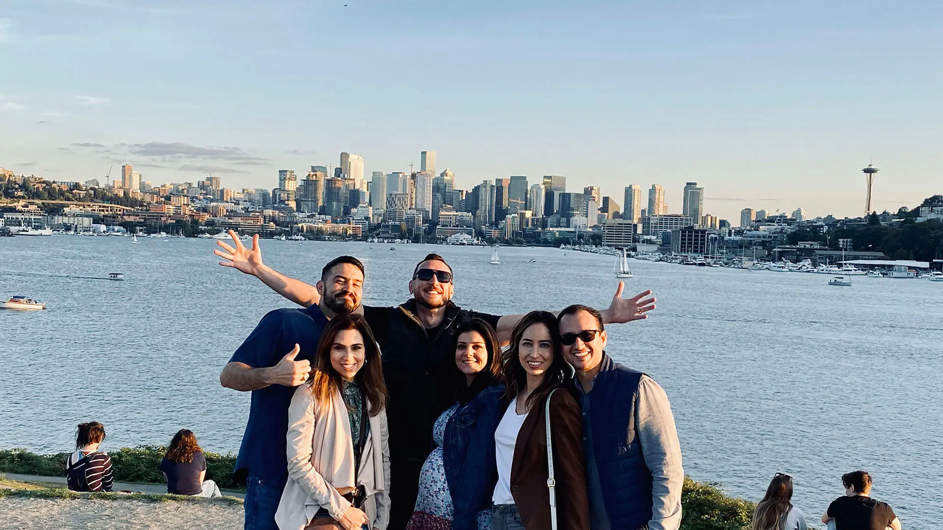 Six people pose and smile in front of a waterfront with a city skyline and the Space Needle in the background during daylight, capturing a perfect moment from their Seattle city tour.