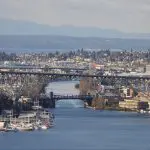 A steel bridge spans a wide waterway in Seattle, with boats docked along the shore and stunning cityscape and mountain sights in the background.