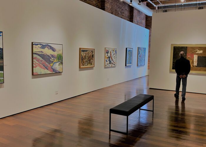 A person stands in an art gallery, part of a private Seattle tour, observing paintings displayed on the walls. A bench is conveniently positioned in the center of the room.